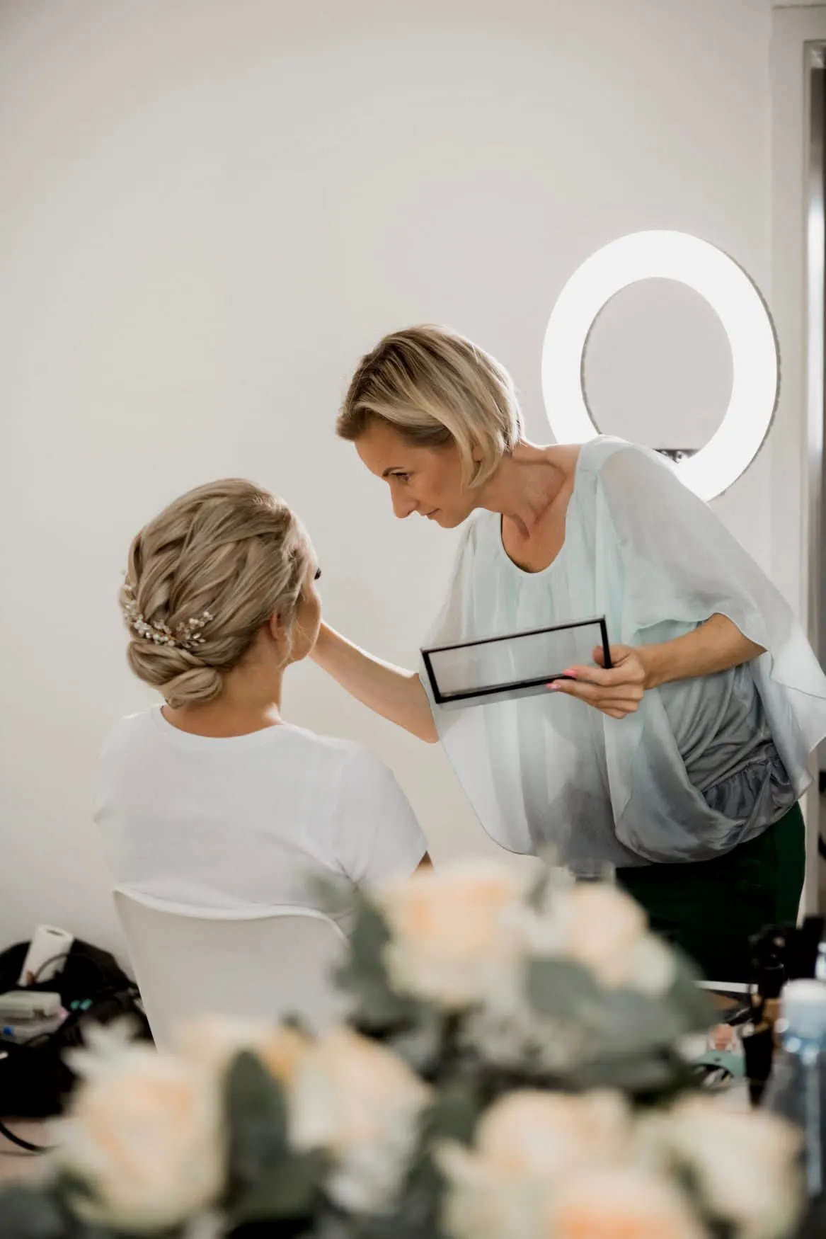 Make-Up und Hairstylistin Artist Iwona Buchna aus Düren schminkt eine sitzende Frau mit gestyltem blonden Haar mit Palette und Pinsel, mit einem Ringlicht im Hintergrund und einem verschwommenen Blumenstrauß im Vordergrund.