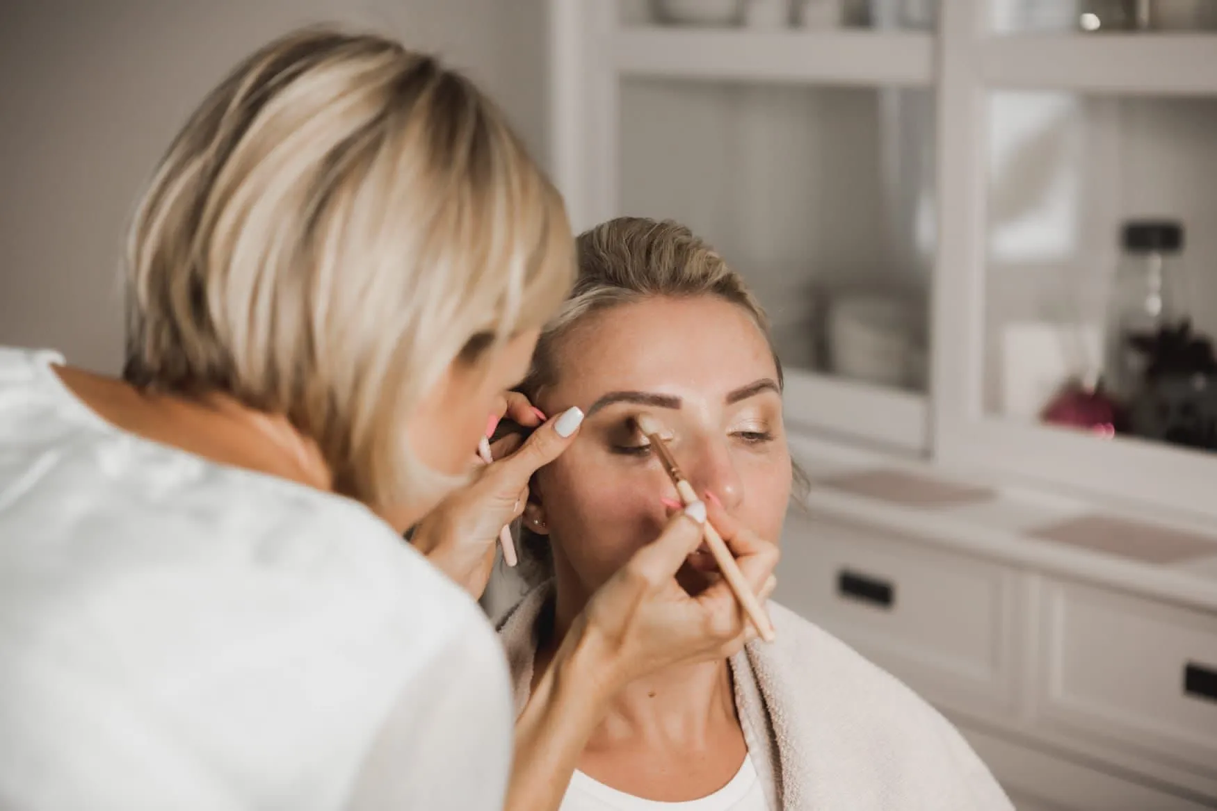 Iwona Buchna - Make-Up Artist für Fotoshootings, Videodrehs und Fashionshows - trägt Lidschatten auf eine Frau mit blondem Haar auf, die in einem weißen Raum sitzt. Dabei konzentriert sie sich mit einem Schminkpinsel sorgfältig auf das Augenlid.