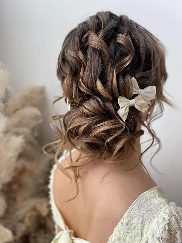 Eine Braut aus Köln mit gewelltem braunem Haar, das von einer geschickten Hairstylistin zu einer eleganten Hochsteckfrisur frisiert und mit einer cremefarbenen Schleife verziert wurde. Sie trägt ein weißes Spitzentop, und der sanft verschwommene Hintergrund ist in neutralen Tönen gehalten. Ein rundum passendes Brautstyling.