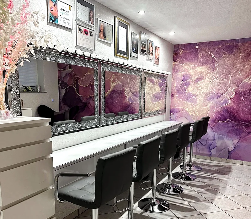 Moderner Schönheitssalon in Düren mit fünf schwarzen Stühlen an einem langen weißen Tresen, perfekt für Make-up-Künstler oder Make-up-Workshops. Mit einer lila und goldenen Marmor-Akzentwand, großen Spiegeln und gerahmten Zertifikaten über dem Tresen.
