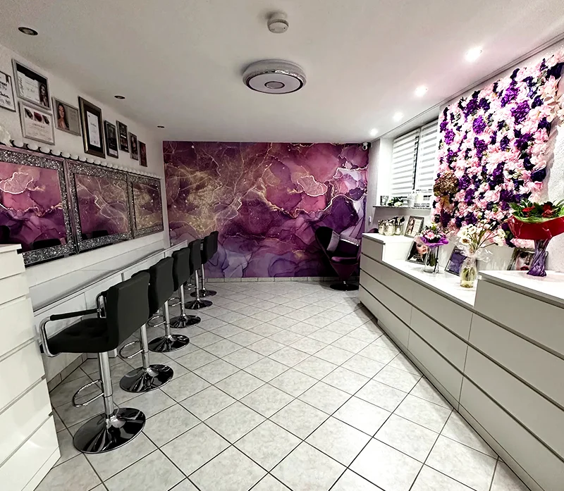 Ein moderner Salon in Düren mit schwarzen Stühlen an verspiegelten Arbeitsplätzen, weißem Fliesenboden und einer leuchtend violetten Akzentwand aus Marmor. Auf der rechten Seite ist eine Wand mit Kunstblumen und Vasen mit Blumenarrangements zusehen.