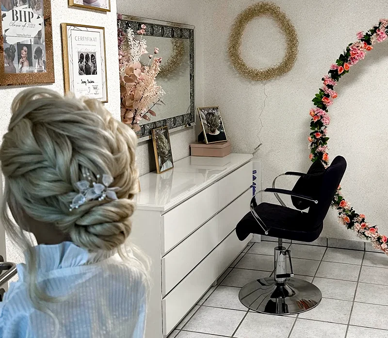Eine Frau mit einer komplizierten blonden Hochsteckfrisur steht in einem Salon, gestylt von einer talentierten Make-up-Künstlerin. Der Salon verfügt über eine weiße Kommode mit gerahmten Fotos und Blumen, einen Wandspiegel, einen schwarzen Stuhl und Blumen- und Kranzdekorationen an den Wänden.
